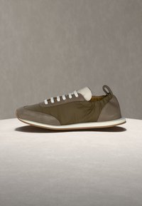 Χαμηλού προφίλ αθλητικό παπούτσι με ελαιόχρωμο ύφασμα, καφέ σουέντ λεπτομέρειες, λευκά κορδόνια, θηλιά στο πίσω μέρος και λευκή σόλα πάνω σε ουδέτερη επιφάνεια.