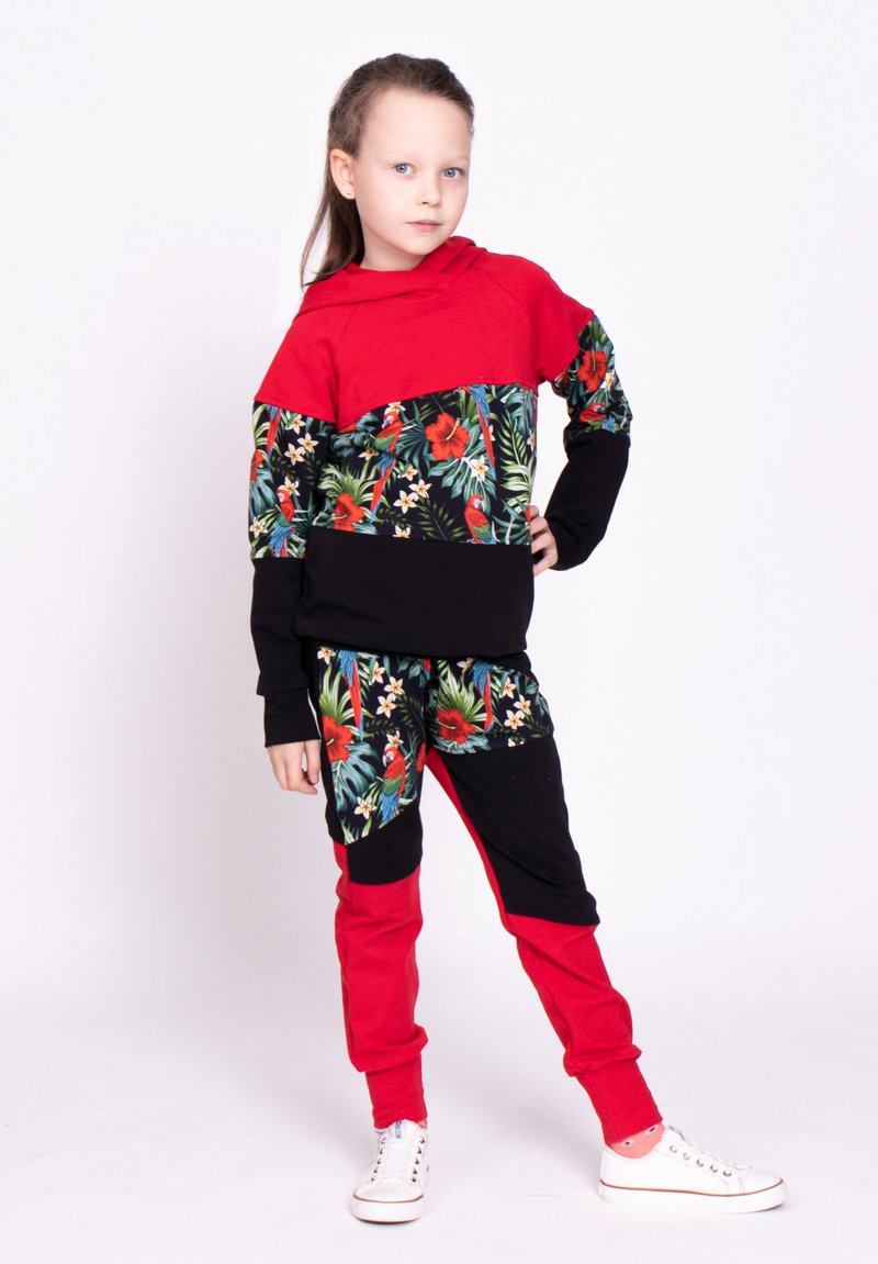 Czerwona i czarna bluza z kapturem w kwiatowy wzór; pasujące spodnie; mieszanka bawełny; wyróżnia się intensywnymi kolorami i graficznymi nadrukami. Białe buty sportowe.