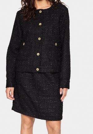 Femme portant une veste texturée noire boutonnée avec des boutons dorés et une jupe assortie, debout devant un fond clair uni.