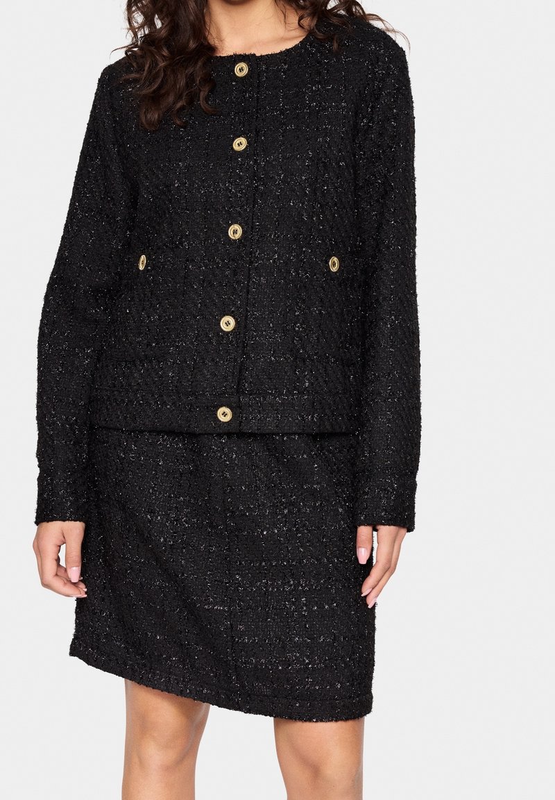 Femme portant une veste texturée noire boutonnée avec des boutons dorés et une jupe assortie, debout devant un fond clair uni.