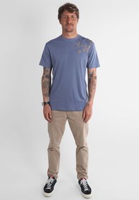 Homme tatoué portant un T-shirt bleu avec un motif floral à l'épaule, un pantalon beige, des baskets noires et une montre, se tenant devant un fond blanc.