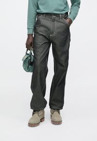 Pantalones vaqueros de mezclilla gris oscuro con costuras blancas, con un diseño de pierna recta. Combinados con botas verde claro y una pequeña bolsa verde.