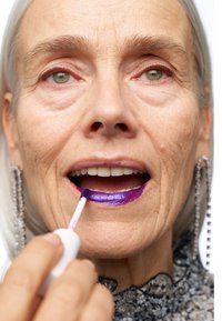 Un rouge à lèvres liquide violet est appliqué avec un applicateur. Les lèvres sont tracées avec du violet profond et le mannequin porte des boucles d'oreilles en argent. La peau est claire.