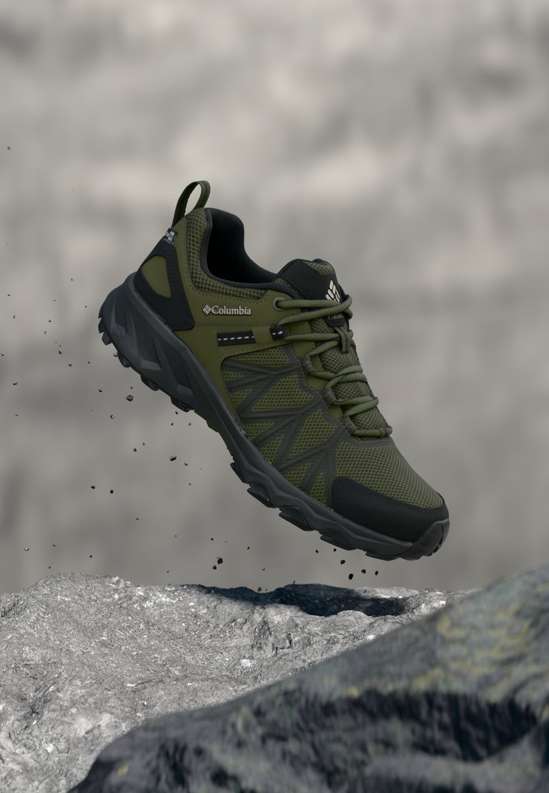 Πράσινο ελιάς και μαύρο αναρριχητικό παπούτσι Columbia κρεμασμένο πάνω από βρ αχώδες έδαφος με σωματίδια χώματος να πέφτουν, σε θολό γκρι φόντο.