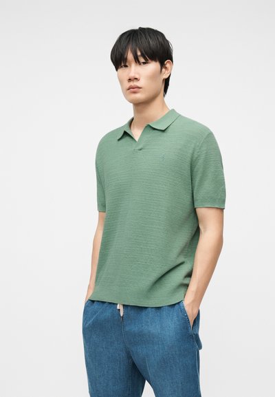 Νεαρός άνδρας που φοράει ένα υφασμάτινο πράσινο πόλο με κοντά μανίκια και μπλε παντελόνι με κορδόνι στη μέση, στέκεται με τα χέρια στις τσέπες μπροστά σε ένα λευκό φόντο.