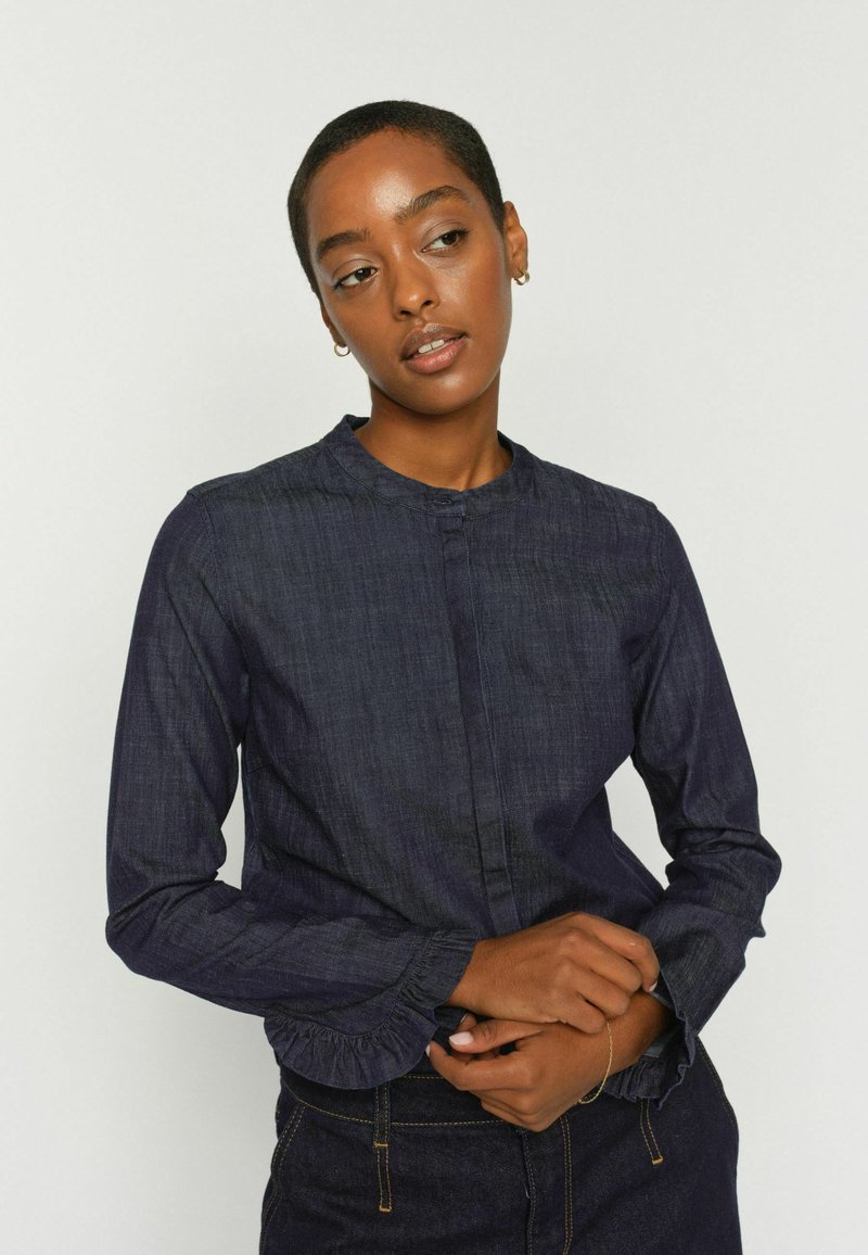 Mladá žena s krátkymi vlasmi, oblečená v tmavej rifľovej dlhorukávej košeli, so zloženými rukami, mierne otočená doľava, na jednoduchom pozadí.