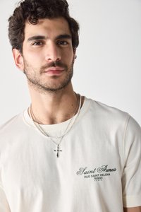 T-shirt en coton beige avec du texte vert, col rond et manches courtes. Colliers en chaîne argentée en superposition avec un pendentif en forme de croix.