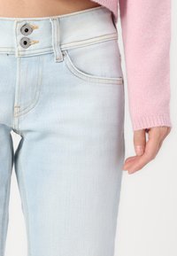 Ανοιχτό μπλε τζιν παντελόνι με ψηλή μέση, κλείσιμο με δύο κουμπιά, πέντε τσέπες και διακριτές ραφές. Υφή ελαφρώς φθαρμένη.