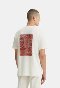 T-shirt bianco a maniche corte con stampa grafica rossa sulla schiena, che rappresenta un paesaggio urbano con edifici, alberi e un'auto.