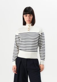 Donna con capelli lisci scuri, che indossa un maglione a righe bianche e blu navy con bottoni dorati e pantaloni neri larghi, in piedi contro uno sfondo bianco.