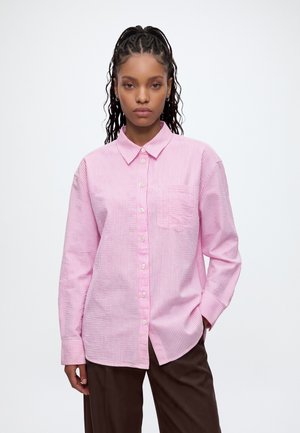 Jeune femme aux cheveux tressés portant une chemise rose et blanche rayée à boutons et un pantalon marron foncé, debout avec une main dans la poche.