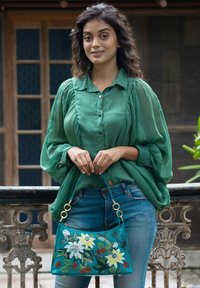 Teal handtas met bloemenprint, gouden hardware en een zachte textuur. Model draagt een groene blouse met pofmouwen en denim jeans.