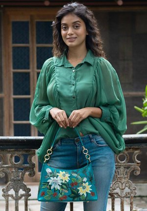 Teal handbag with floral print, gold hardware, and a soft texture. Model wears a green blouse with puffed sleeves and denim jeans.