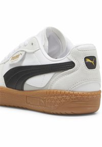 Valkoiset lenkkarit mustilla samettikoristeilla, joissa on kumipohja, teksturoitu kantapää ja kultainen PUMA-logo sivulla ja kielessä.