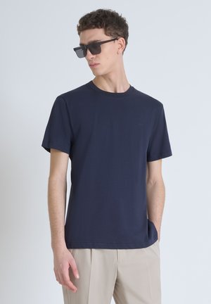Camiseta de algodón azul marino con mangas cortas y cuello redondo acanalado. Presenta un sutil detalle del logo en el pecho, combinada con pantalones de un beige claro.
