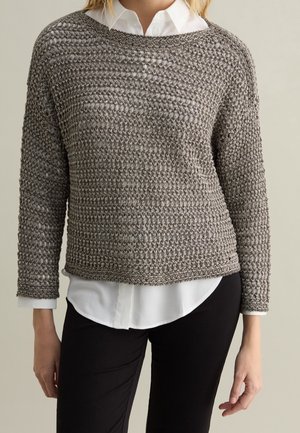 Vrouw die een grijze, losgebreide trui draagt over een wit overhemd met knopen, gecombineerd met een zwarte broek, tegen een effen achtergrond.