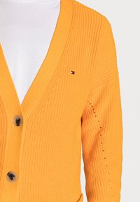 Neulecardigan kirkkaan oranssina, jossa on V-pääntie, kaksi nappia ja pieni logo vasemmassa rinnassa. Rakennekangas ja väljä malli.