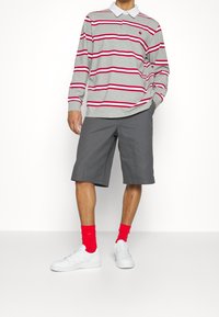 Graues Langarm-Polohemd mit roten und weißen Streifen, kombiniert mit grauen Shorts. Trägt rote Socken und weiße Turnschuhe.