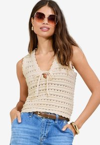 Beige gehäkeltes Oberteil mit V-Ausschnitt, das gewellte Kanten und eine Bindeschnur vorne hat. Kombiniert mit blauen hochgeschnittenen Jeans und einem braunen Gürtel.