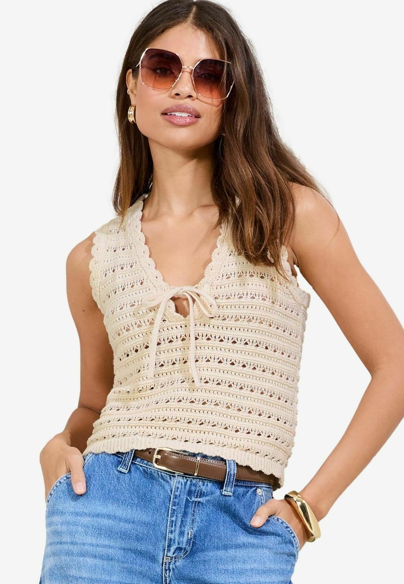 Beige gehäkeltes Oberteil mit V-Ausschnitt, das gewellte Kanten und eine Bindeschnur vorne hat. Kombiniert mit blauen hochgeschnittenen Jeans und einem braunen Gürtel.