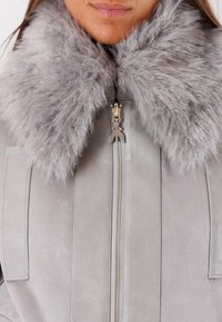 Donna che indossa una giacca in suede grigio chiaro con un ampio colletto in pelliccia soffice e una zip centrale argentata, mostrando le labbra e la parte inferiore del viso.