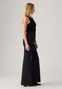 Vrouw met lang blond haar die zijwaarts staat, gekleed in een zwarte mouwloze top en hoge taille zwarte wijde jeans met zwarte schoenen.