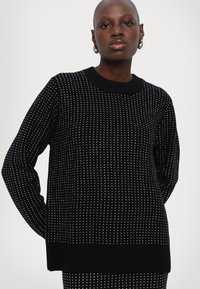 Pull noir en tricot avec un motif texturé de petits pois blancs, doté d'un col rond et de poignets et d'un ourlet côtelés.