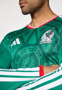 Πράσινη αθλητική φανέλα με έμβλημα του Μεξικού, που διαθέτει περίπλοκα σχέδια, V-λαιμό με κόκκινες λεπτομέρειες και λευκές ρίγες Adidas στα μανίκια.