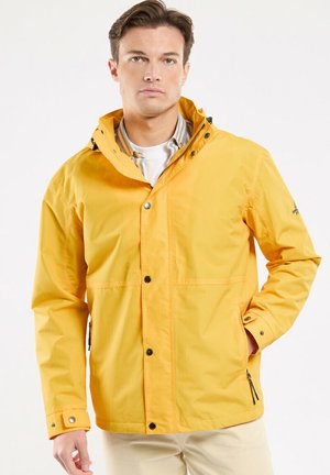 Homme portant une veste à capuche jaune vif par-dessus une chemise rayée, debout avec une main dans la poche sur un fond uni.