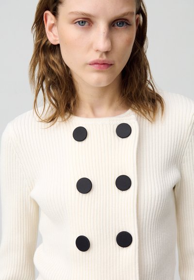 Cardigan a coste crema con colletto rotondo e sei grandi bottoni neri come chiusure, che mette in evidenza un design testurizzato.