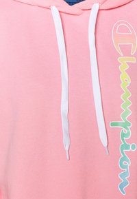 Rosa bomullshoodie med vita dragsko och en flerfärgad "Champion"-logotyp på framsidan i böjd text. Mjuk textur.