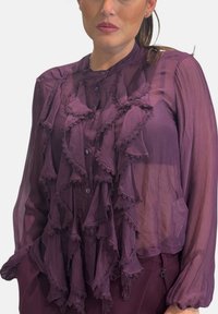 Camicia viola trasparente con dettagli arricciati sul davanti, dotata di chiusura con bottoni e rifinitura in pizzo sottile lungo i bordi.