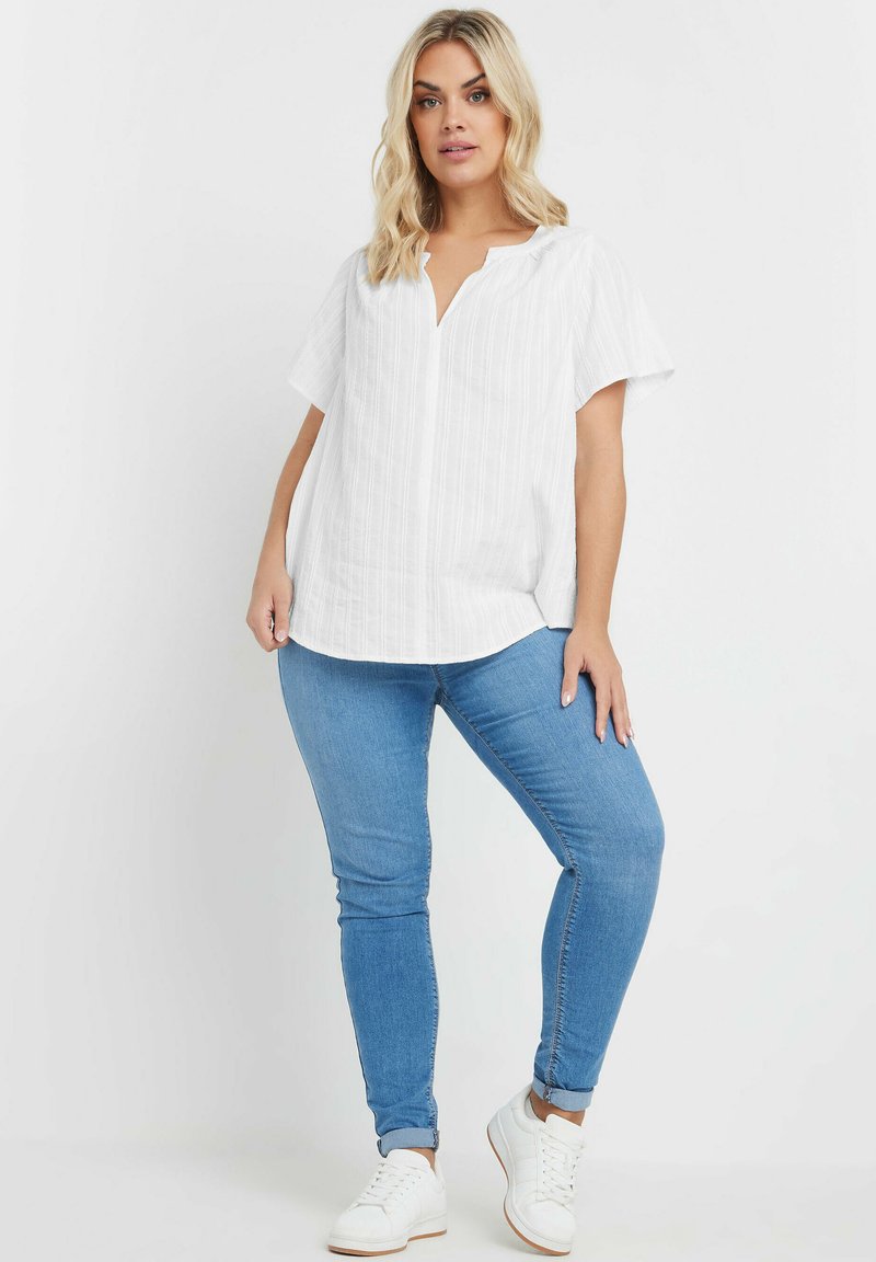 Top bianco a maniche corte con scollatura a bottoni e texture orizzontale. Abbinato a jeans skinny blu chiaro e sneaker bianche.