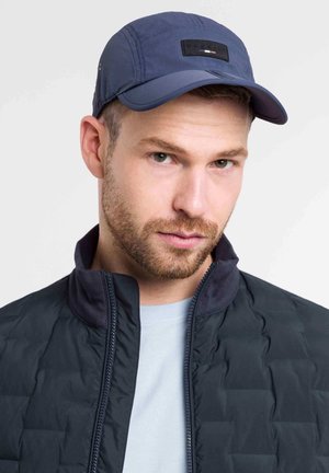 Șapcă de baseball bleumarin, dintr-un material moale, cu o margine curbată și un petic negru pe partea din față. Purtați cu o jachetă neagră matlasată și o cămașă bleu deschis.