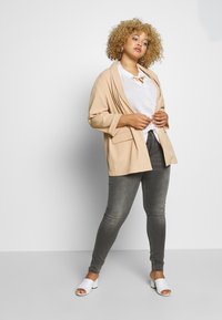 Beige oversized blazer, white textured blouse with a tie, gray skinny jeans, and white open-toe mules. Smooth fabric, relaxed fit.