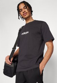 Juodas medvilninis marškinėliai su baltu "adidas" logotipu priekyje, derinami su juodomis kelnėmis ir laikant mažą juodą maišelį su tinkliniais akcentais.