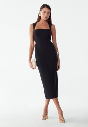 Femme debout dans une robe midi noire sans manches avec un décolleté carré, tenant une pochette blanche, portant des sandales dorées à lanières et un bracelet manchette.