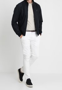 Mężczyzna w czarnej kurtce, jasnoszarym t-shircie, białych spodniach, brązowym pasku i czarnych trampkach, stoi z rękami w kieszeniach.