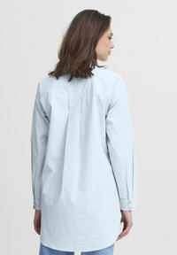 Camisa azul clara con un pliegue en la espalda, mangas largas y puños abotonados, con un corte holgado y una tela lisa y texturizada.