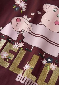 Mörk röd tyg med en tecknad design som visar rosa grisar, blommönster och glittrande guldtext som stavar "HEJ BUMP."