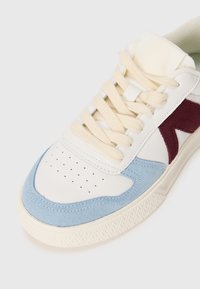 White sneaker with blue and burgundy accents, smooth leather, suede panels, round toe, and textured rubber sole. Cream laces complete the design.
