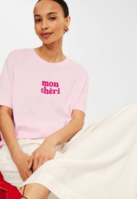 Rosa bomullströja med texten "mon chéri" i fet magenta. Matchad med en vit, flödande kjol som visar en avslappnad passform och enkel design.