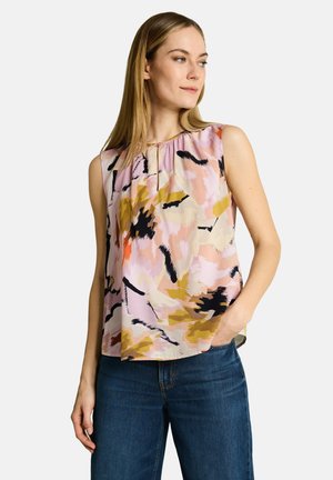 Frau trägt ärmellose Bluse mit pastellfarbenem abstraktem Muster und blaue Jeans, steht mit einer Hand in der Tasche, schaut nach rechts.