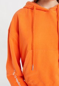 Sweatshirt à capuche orange avec une poche avant, en tissu texturé et cordons avec embouts en métal ; impression du logo en blanc sur une manche.
