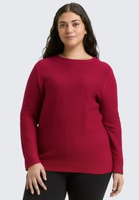 Roter strukturierter Pullover mit langen Ärmeln und einem weiten Ausschnitt, der ein Rautenmuster aufweist. Über schwarzen Hosen getragen.