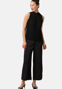 Svart veckad ärmlös topp med hög halsringning, tillsammans med vida svarta byxor och svarta klackskor, mot en ljus bakgrund.