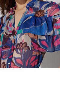 Blouse à motif floral avec des manches volantes, dans des tons de bleu, rose et marron. Fabriquée en tissu léger et légèrement transparent avec des boutons.
