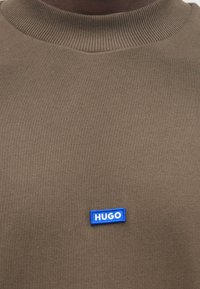 Maglione kaki con collo girocollo a coste e toppa con logo "HUGO" blu. Tessuto liscio, design aderente, cuciture visibili lungo il collo.