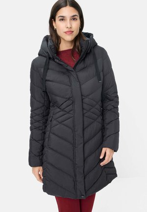 Frau trägt einen schwarzen gesteppten Wintermantel mit Kapuze über einem burgunderroten Pullover und Hose, steht vor einem einfarbigen Hintergrund.