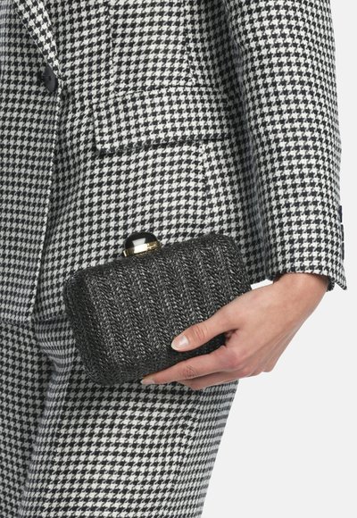 Črno zlata teksturirana torbica s pravokotno obliko in zaobljenim zapiralom, ki jo roka drži ob črno-beli obleki s houndstooth vzorcem.